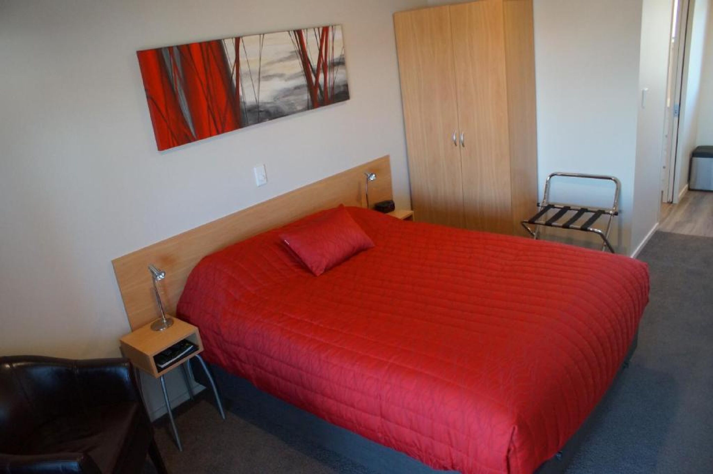 A hotel room with a large bed, a wooden wardrobe, a small table with a lamp, and a chair.