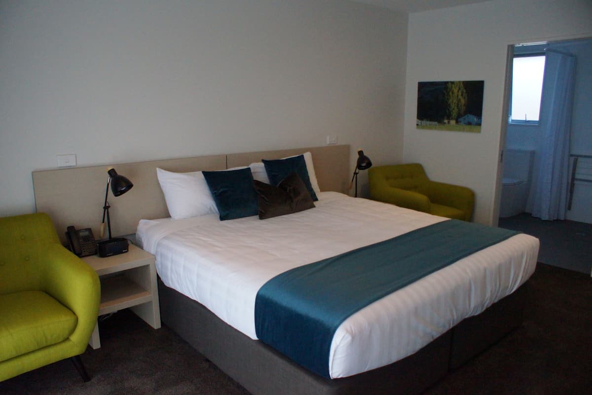 A hotel room with a large bed, two bedside tables, a nightstand, a green armchair, and a painting on the wall.