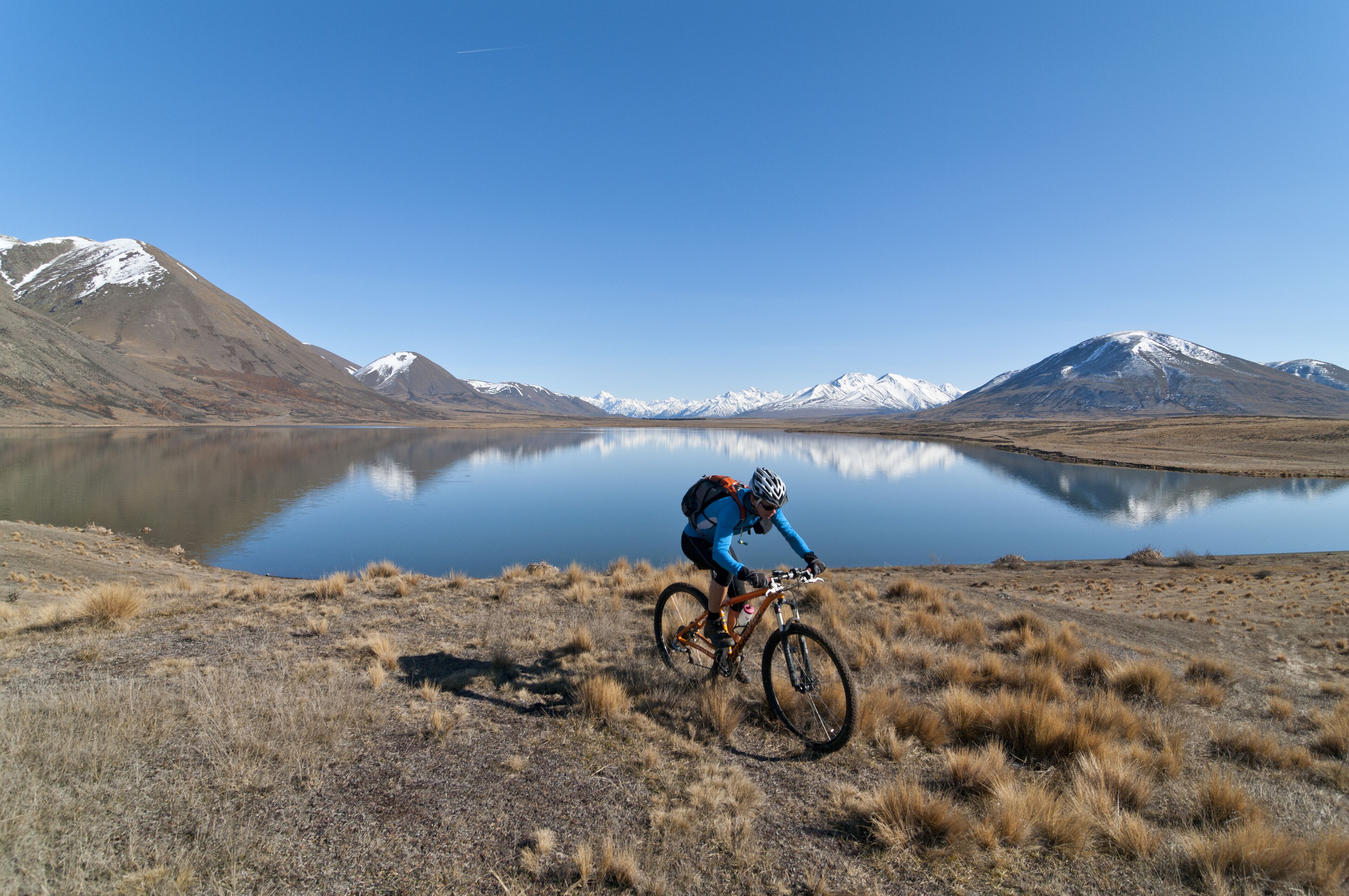 A mountain biker on a trail with a lake and snow-capped mountains in the background.