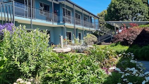 Un edificio de dos pisos con un balcón, rodeado de vegetación exuberante y plantas en flor.
