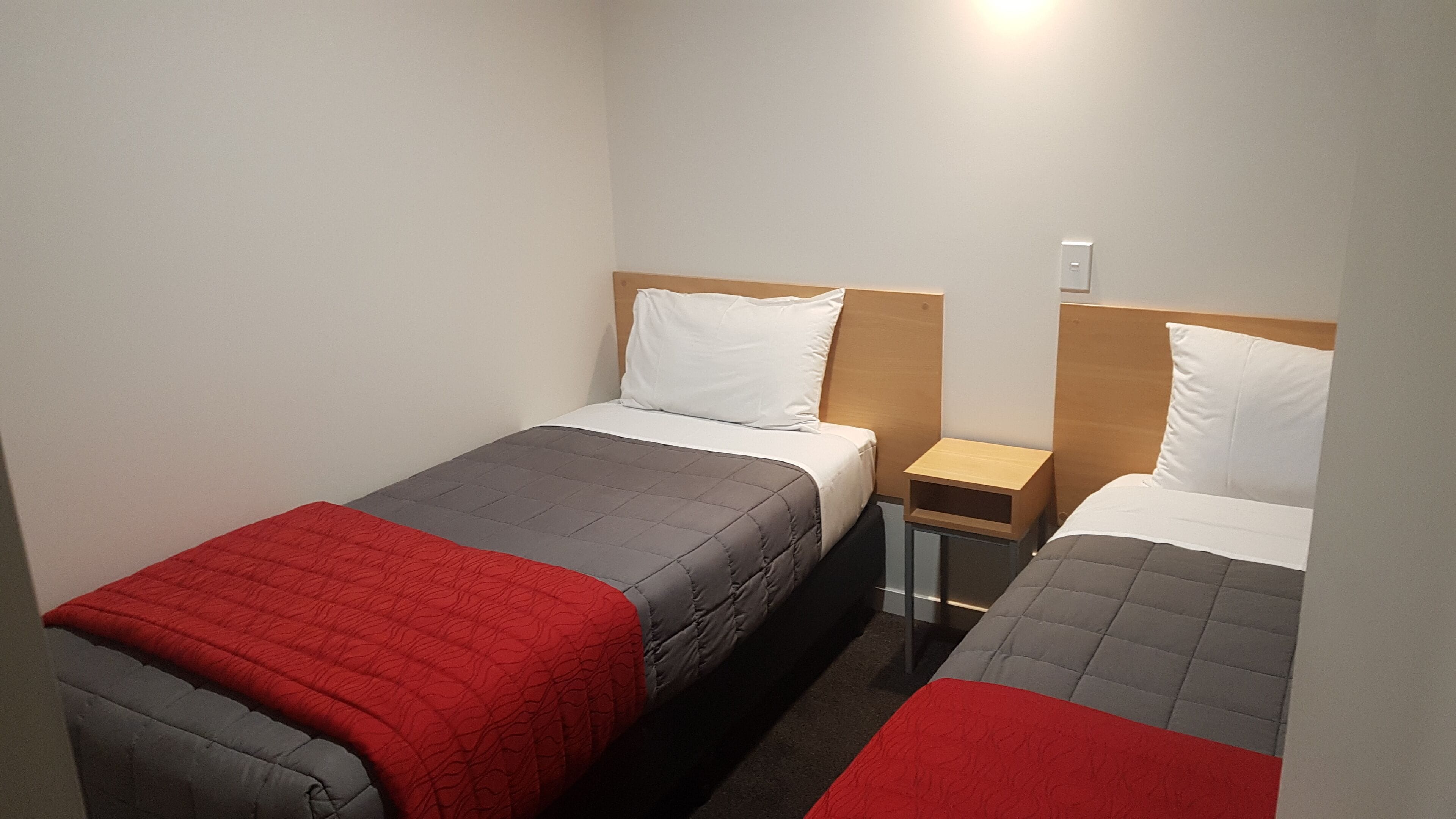 A twin room with two single beds, each with a red and grey quilt, white pillows, and wooden headboards. There is a small wooden nightstand between the beds.