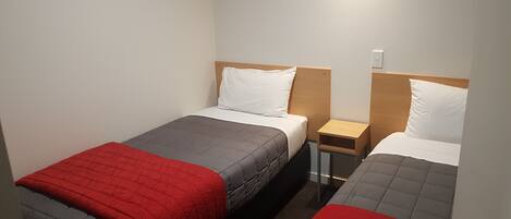 A twin room with two single beds, each with a red and grey quilt, white pillows, and wooden headboards. There is a small wooden nightstand between the beds.