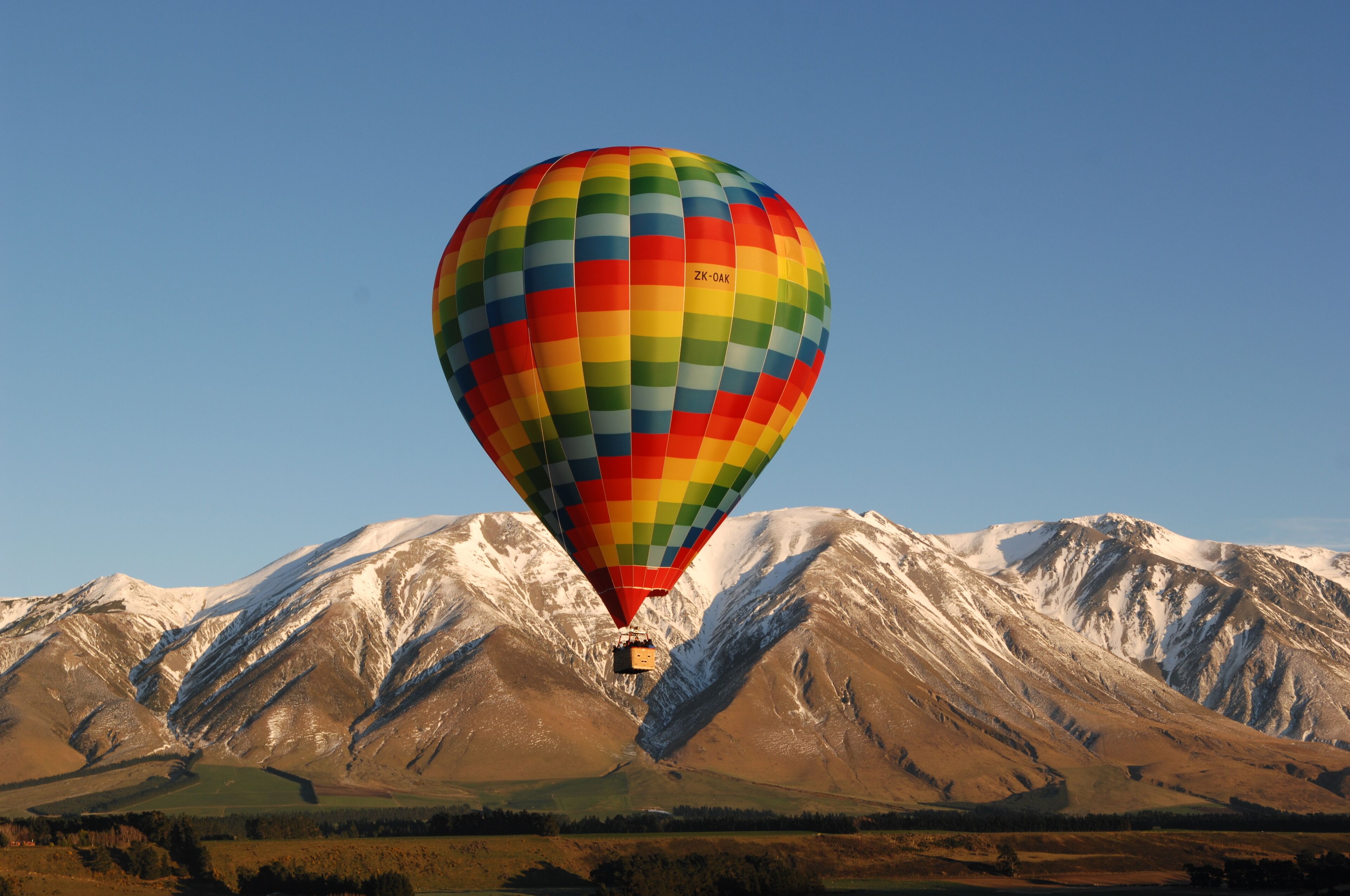 A hot air balloon with a colorful pattern flying over a mountainous landscape with snow-capped peaks.