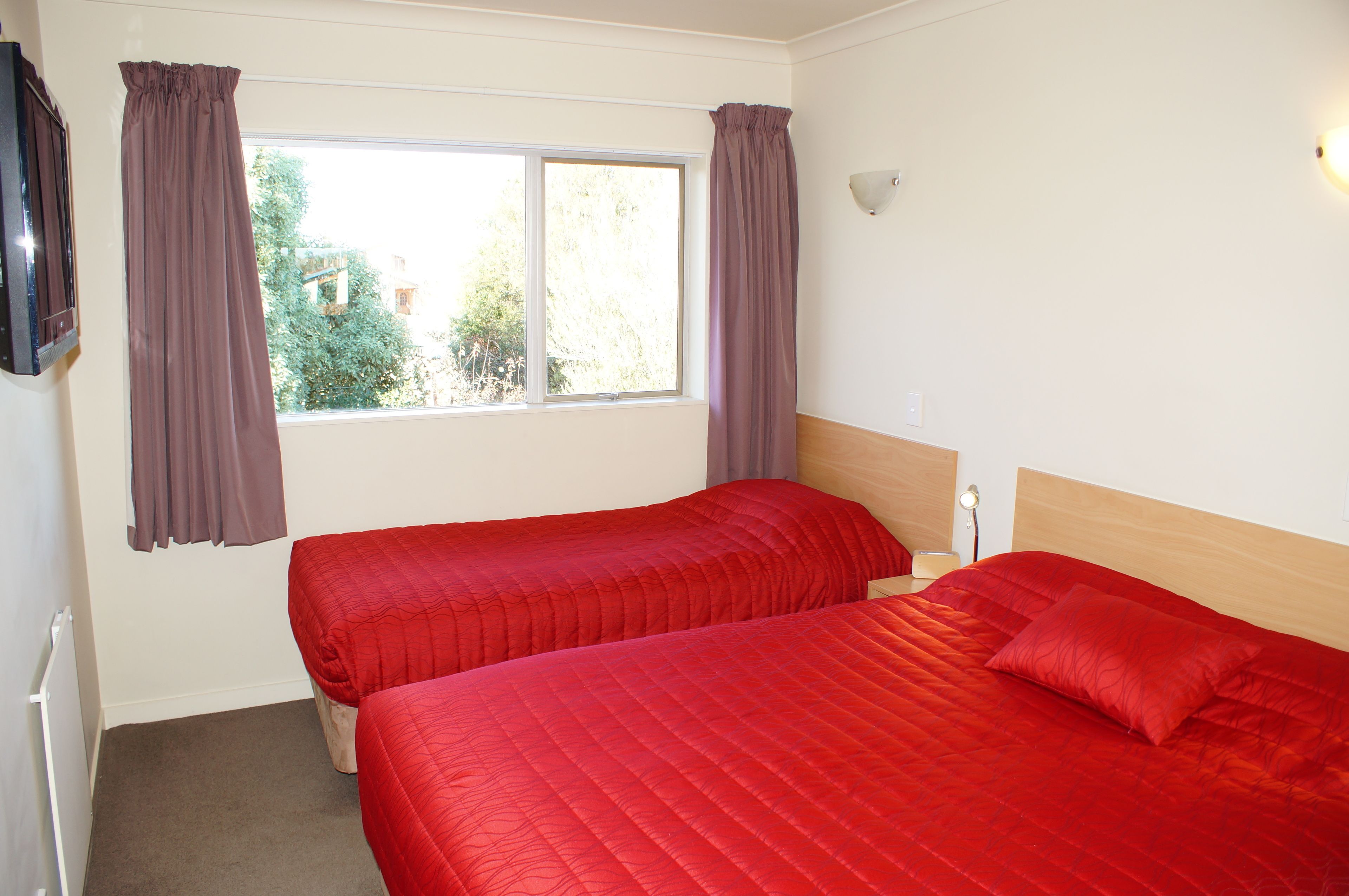 A bedroom with two beds, a window with curtains, a wall-mounted TV, and a bedside lamp.