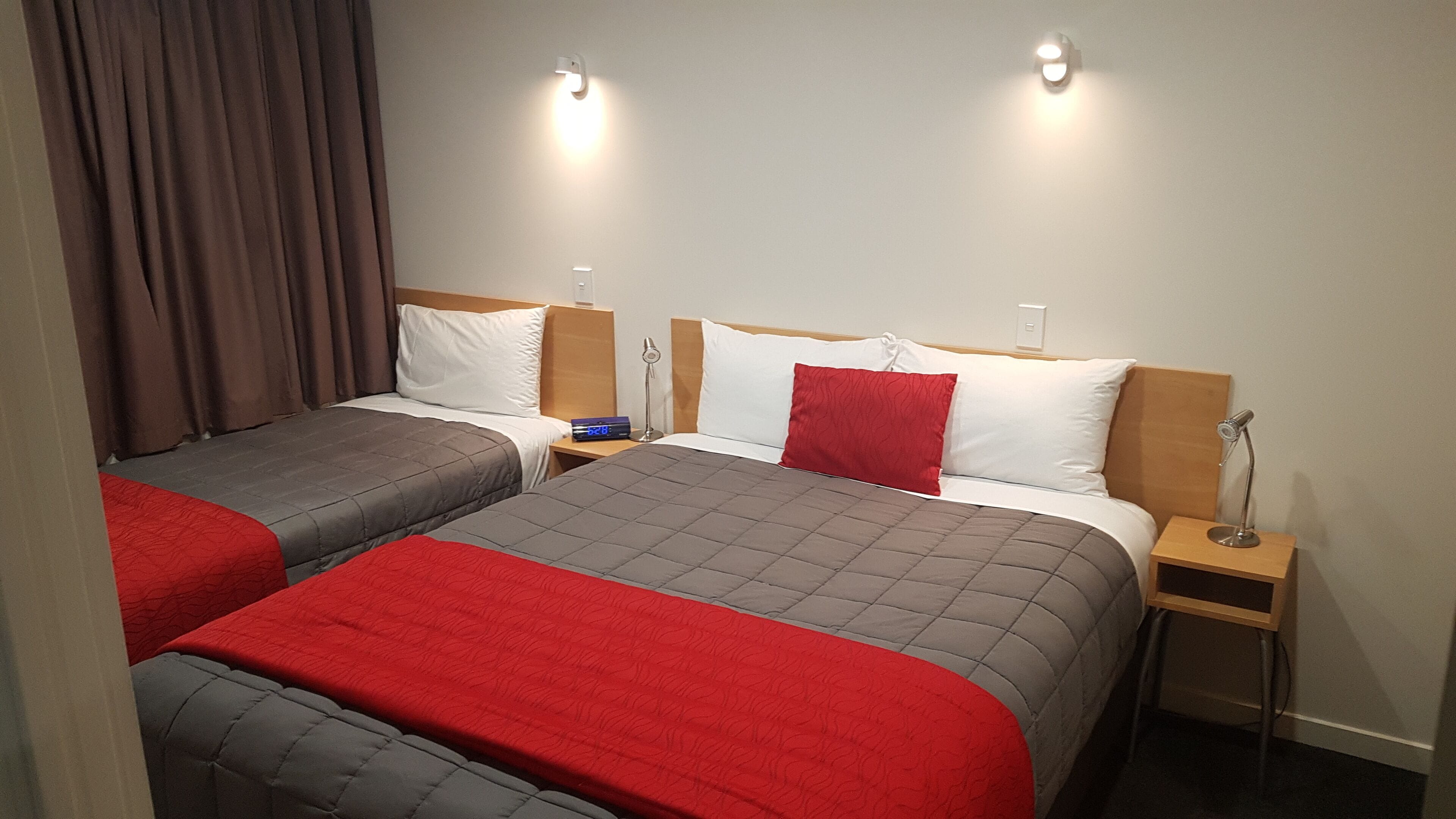 Habitación de hotel con dos camas, cada una con cubrecama rojo y gris, almohadas blancas y cabeceros de madera. Se observan mesitas de noche con lámparas y un reloj digital.