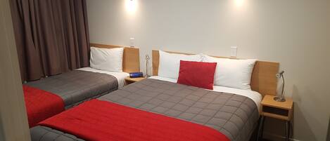 Habitación de hotel con dos camas, cada una con cubrecama rojo y gris, almohadas blancas y cabeceros de madera. Se observan mesitas de noche con lámparas y un reloj digital.
