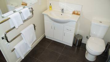 A bathroom with a white sink, mirror, and toilet. There are towels on a rack and a trash can.