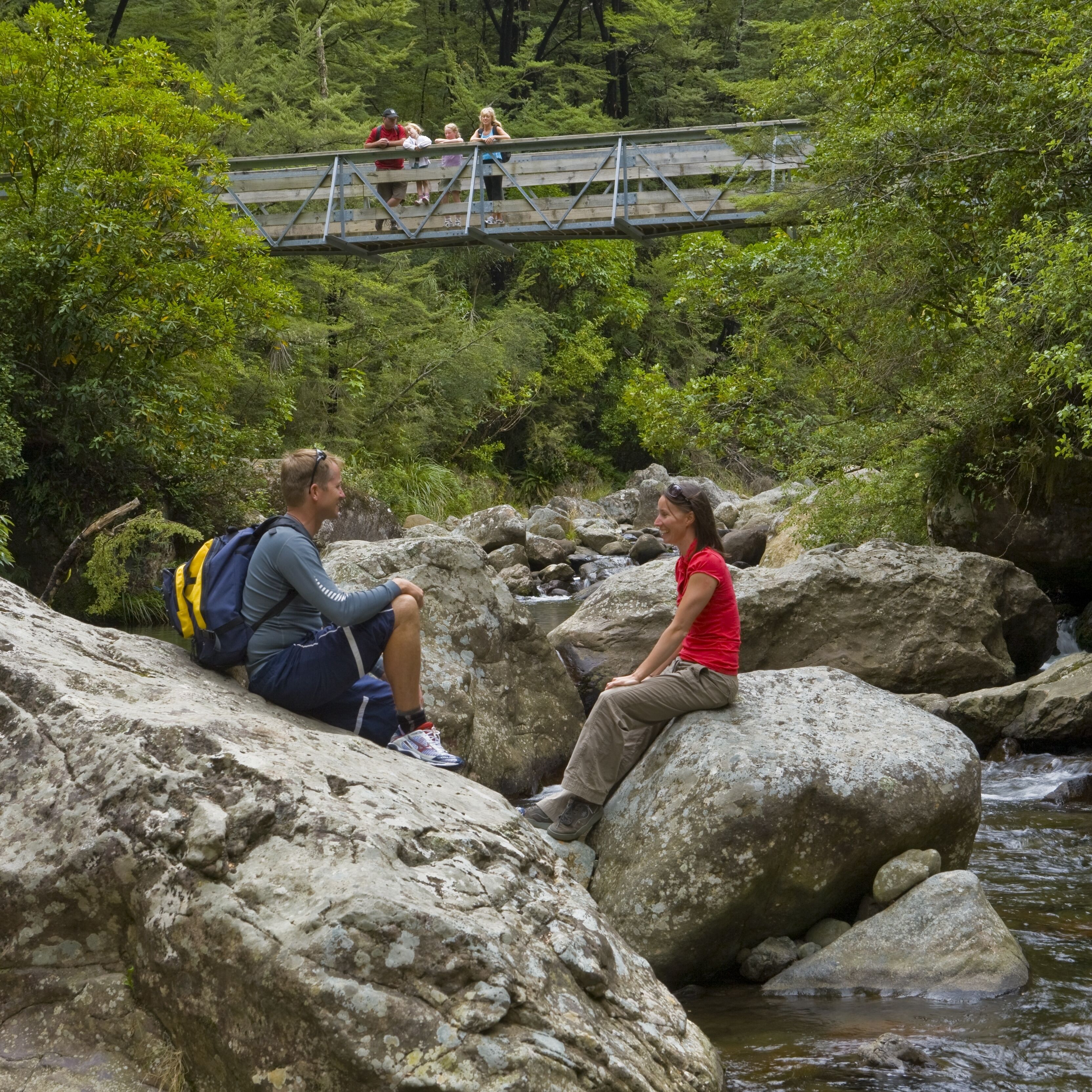 Dos personas sentadas sobre rocas junto a un río, con un puente al fondo.
