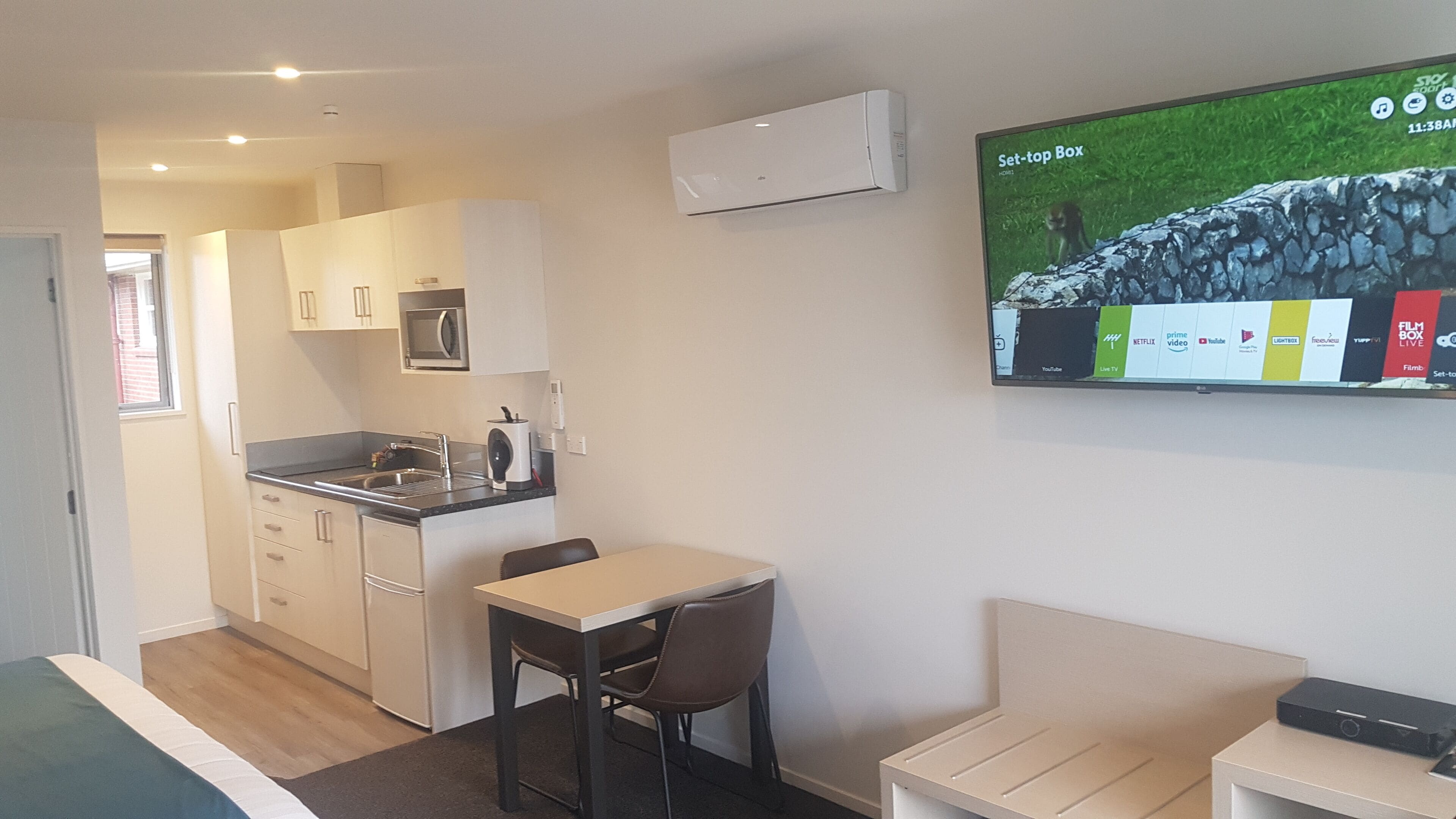 Una cucina compatta con zona pranzo, una TV a parete che mostra l'interfaccia di un servizio di streaming e un piccolo angolo salotto.