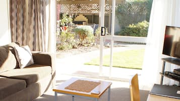 A living room with a sofa, a coffee table, a TV, and a laptop. There is a sliding glass door leading to an outdoor area with a garden.
