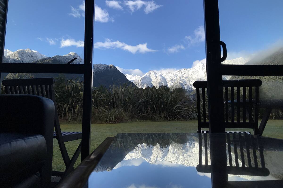 A view from a window with a table and chairs, overlooking snow-capped mountains and a clear sky.