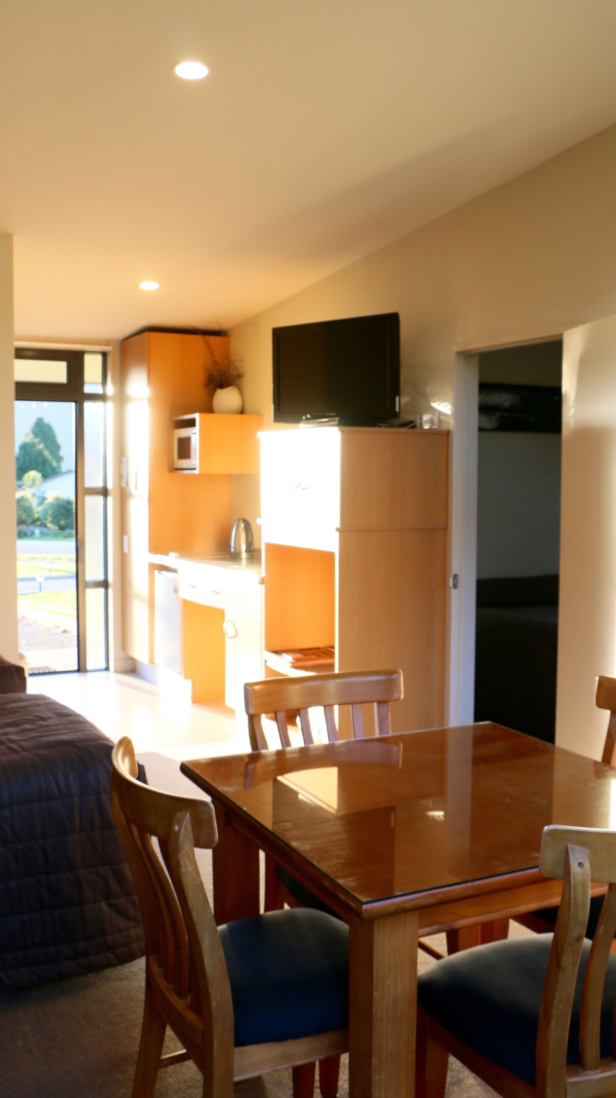 Un comedor con mesa y sillas de madera, una cocina con microondas y armarios, y un televisor montado sobre los armarios.