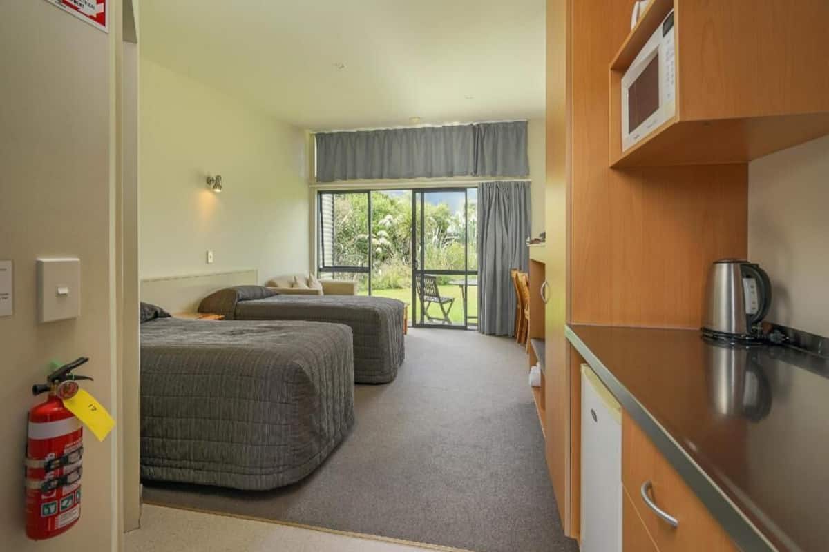 A hotel room with two beds, a kitchenette, a fire extinguisher, and a view of the outdoors.