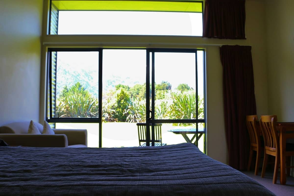 A bedroom with a bed, a chair, a table, and a view of greenery through large windows.