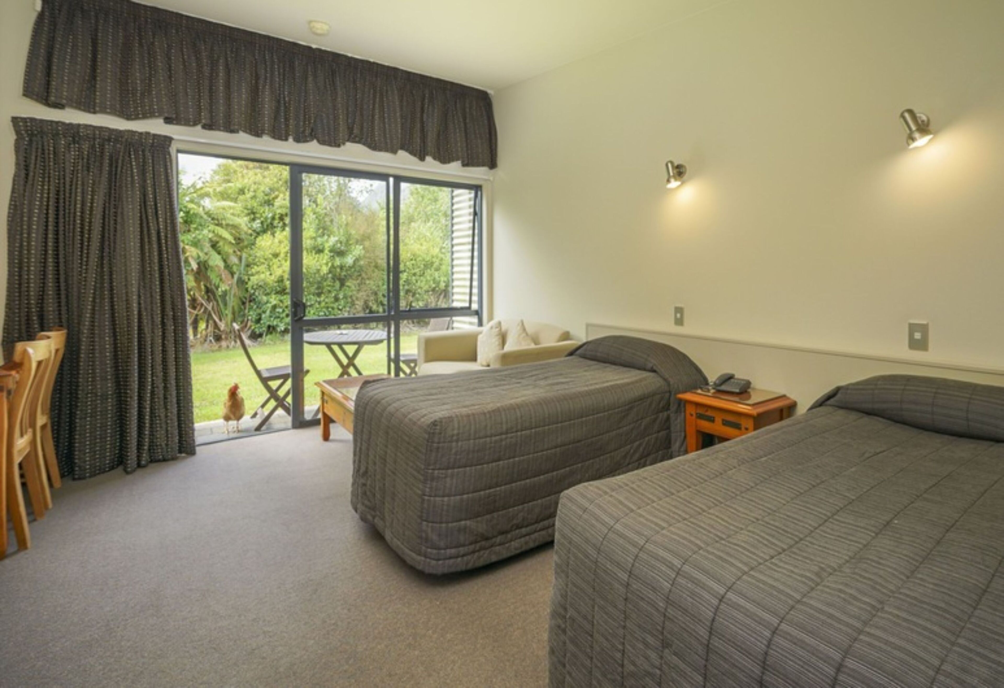 Un dormitorio con dos camas, un escritorio y una silla. Se ve una ventana con cortinas y vistas al exterior con vegetación.