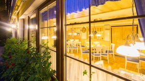 A well-lit interior of a restaurant with multiple dining tables and chairs, large windows, and hanging pendant lights.
