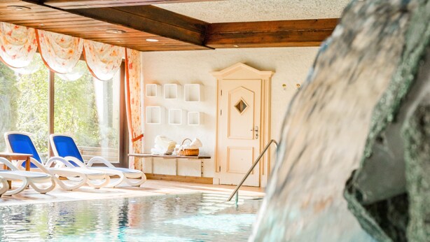 An indoor swimming pool with clear water, wooden beams, and blue lounge chairs.