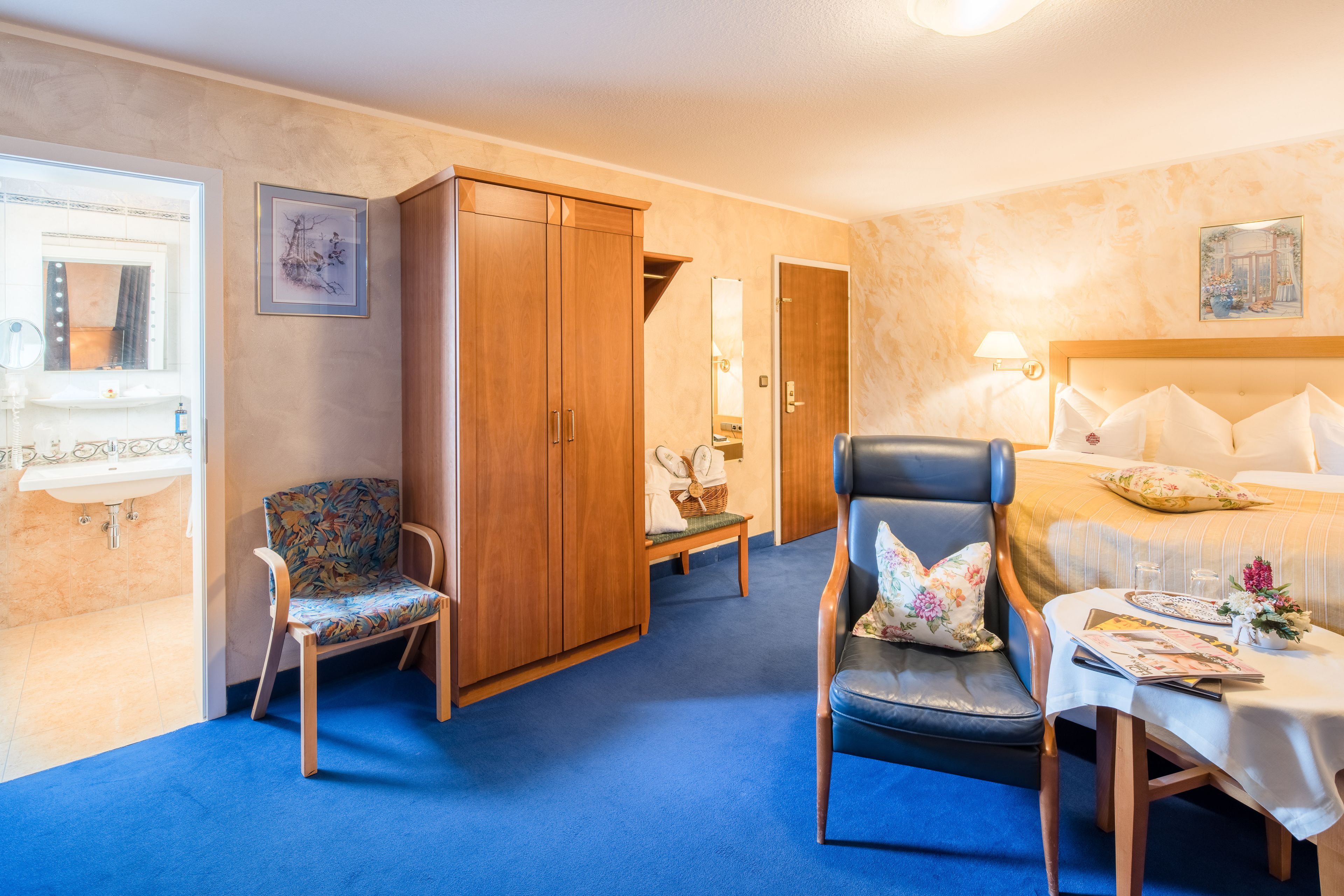 Una habitación de hotel con cama, una silla, una mesa con un jarrón, un armario y un baño con lavamanos y espejo.