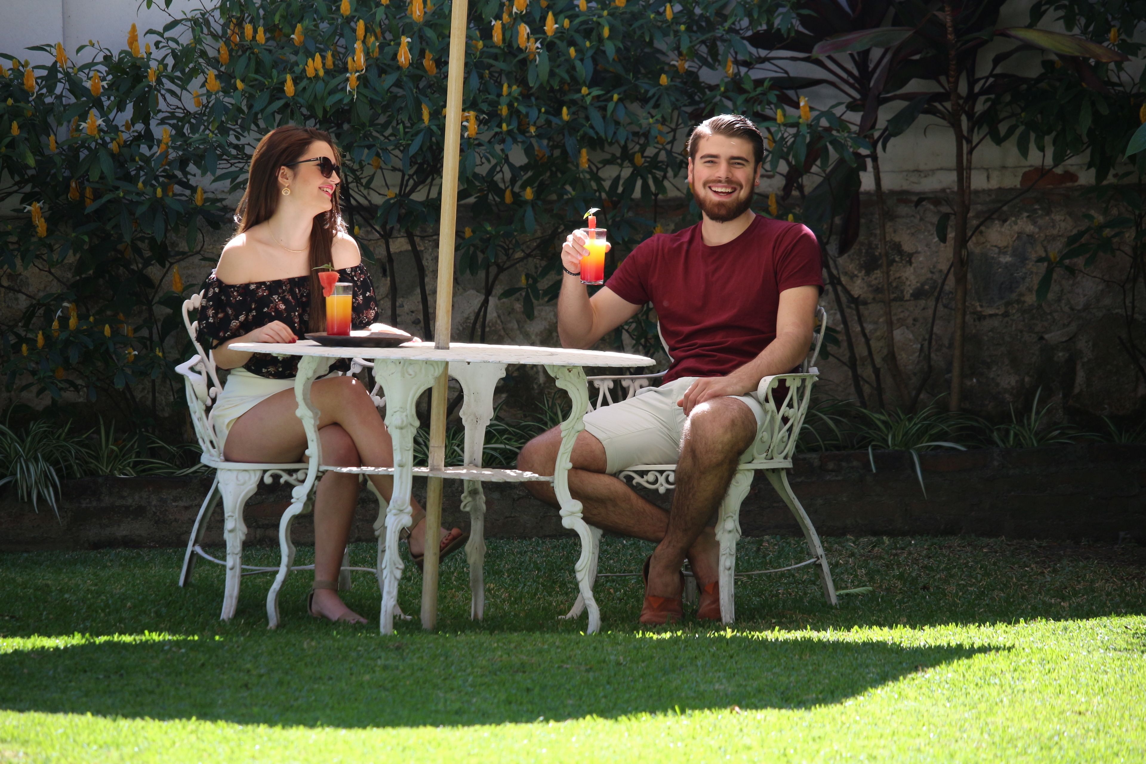 Two people seated at a round table outdoors with a grassy area and plants in the background.