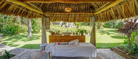 A thatched-roof outdoor spa area with a massage table, a wooden cabinet with plants, and a garden view.