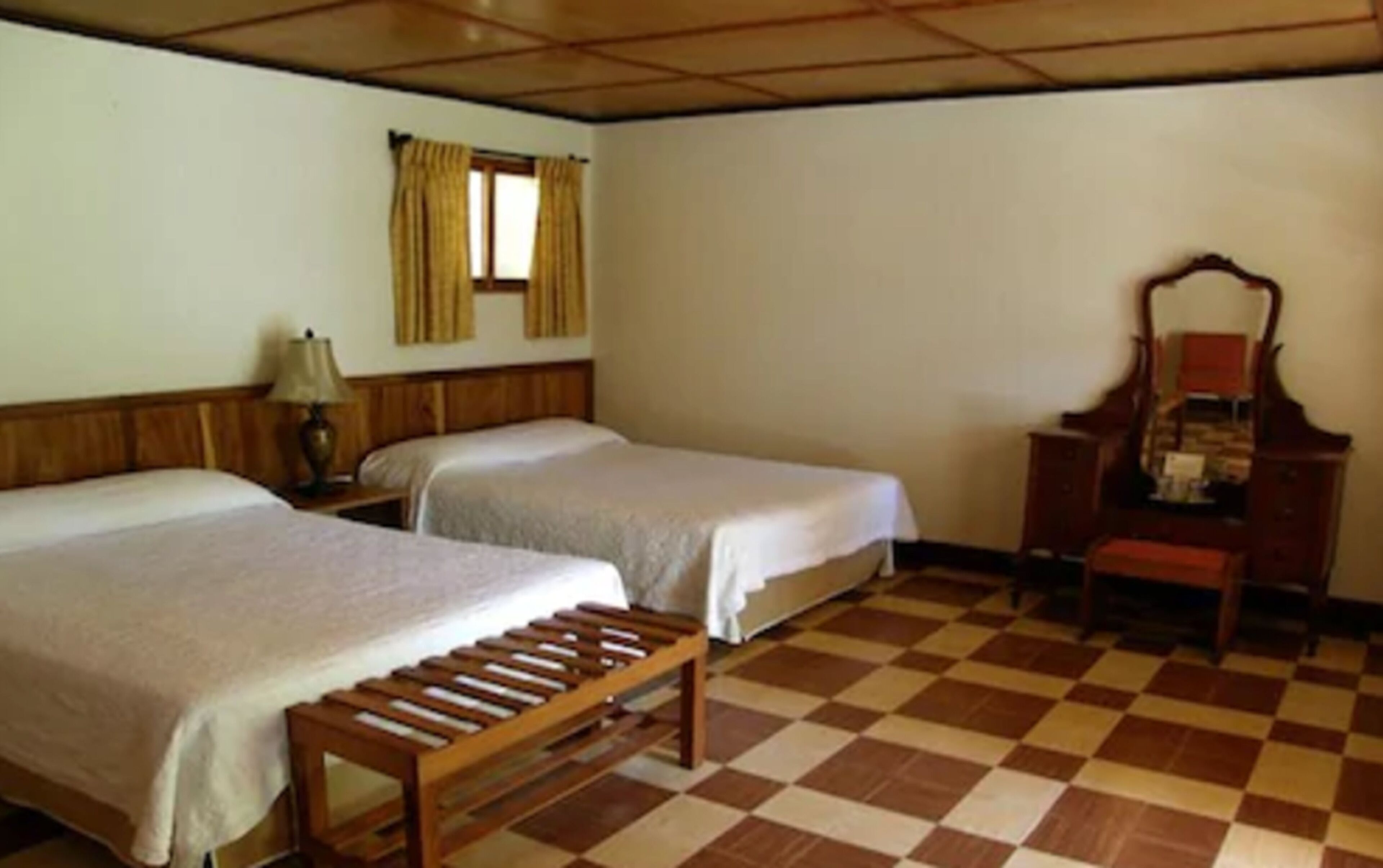 A room with two beds, a wooden bench, a desk with a mirror, and a checkered floor.