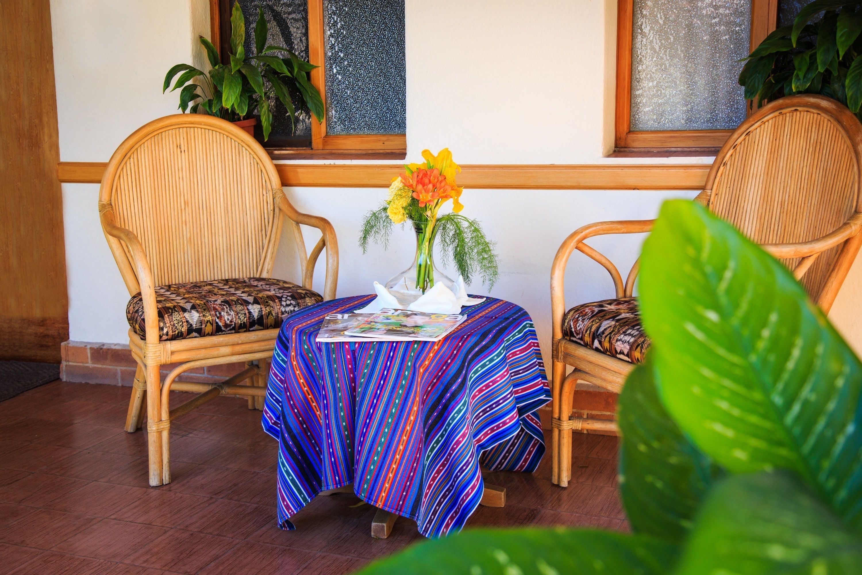 Una habitación con dos sillones de mimbre, una mesita con mantel a rayas de colores y un jarrón con flores.