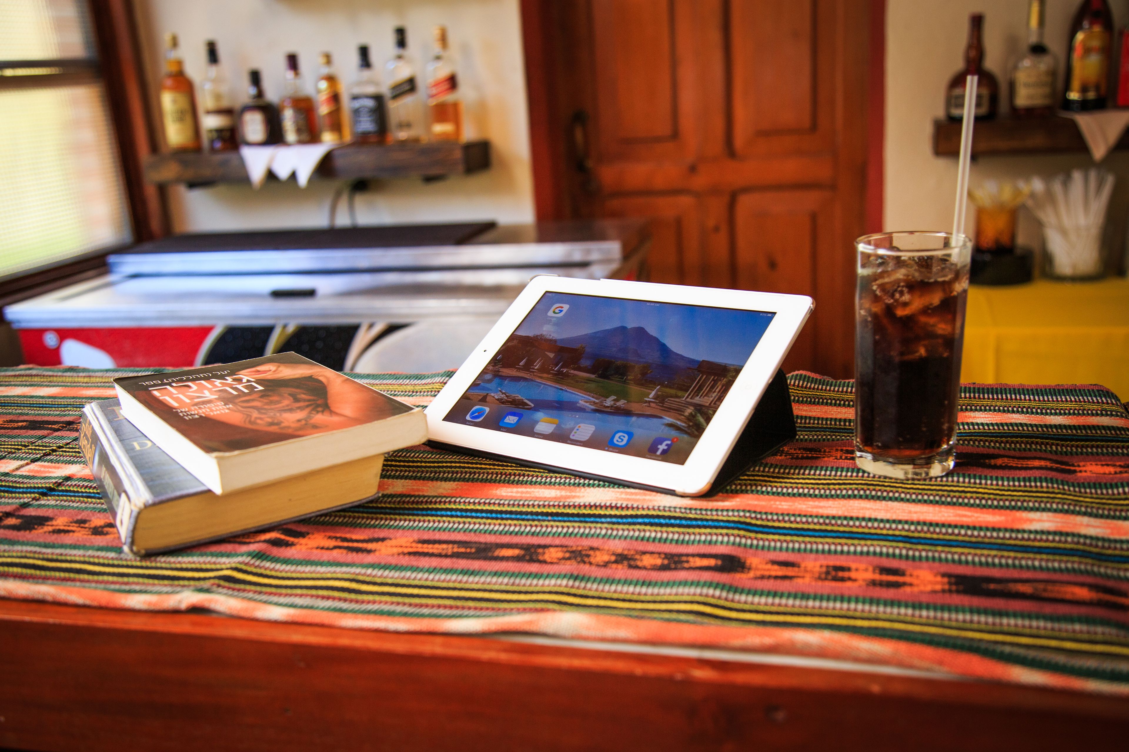A tablet displaying a mountain landscape, a book, and a glass of iced beverage on a patterned tablecloth.