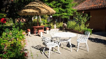 Un patio exterior con una mesa redonda y sillas blancas, rodeado de abundante follaje y un toldo de paja.