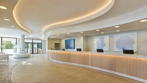 A modern reception area with a curved reception desk, a large flat-screen TV, and framed artwork on the wall. - Torreserena Resort (Ginosa)