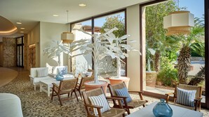 A modern indoor space with a seating area featuring wicker chairs and a white table, large windows offering views of greenery, and a tiled floor. - Torreserena Resort (Ginosa)