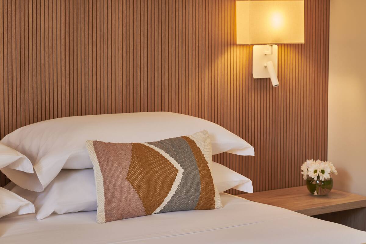 Ein Hotelzimmer mit Bett, Kissen, einem Nachttisch mit Lampe und einer Wandleuchte.
