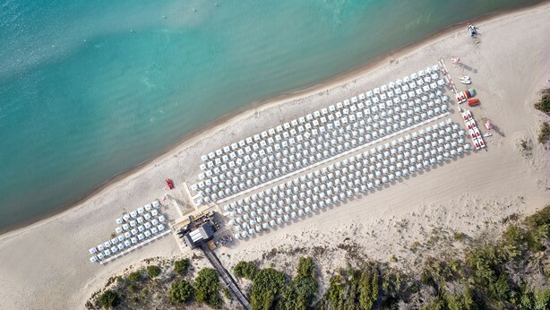 An aerial view of a beach with rows of white beach umbrellas and lounge chairs, a small building, and sparse vegetation. - Torreserena Resort (Ginosa)