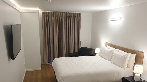 A hotel room with a bed, bedside table, lamp, TV, and a desk with a vase of flowers.