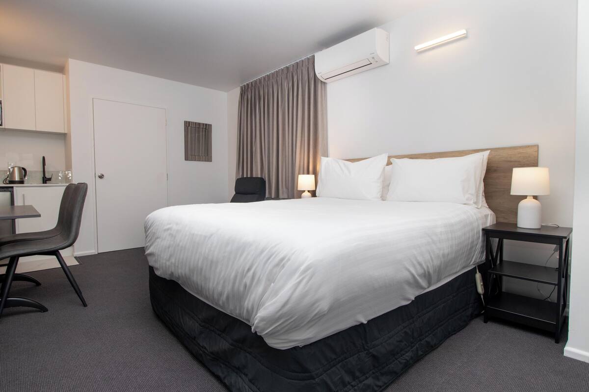 A hotel room with a large bed, a desk with a chair, a small table with a lamp, and a wall-mounted air conditioner.