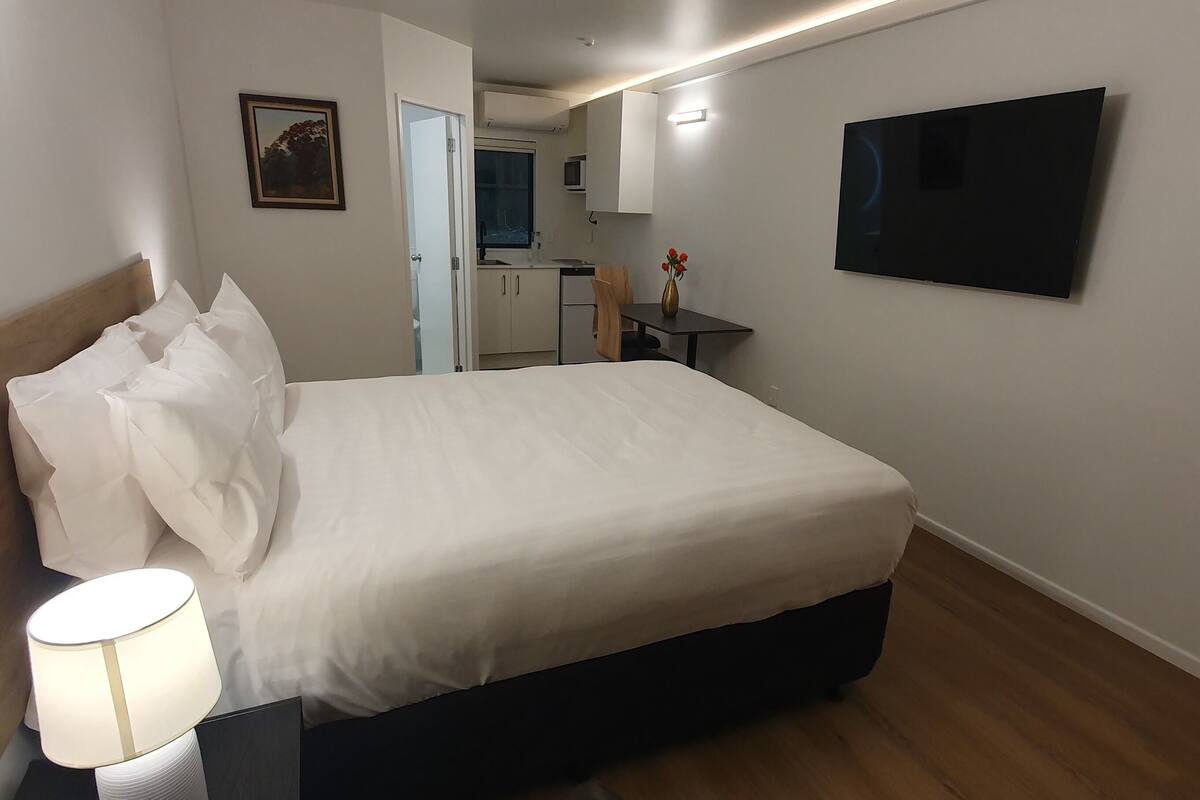 A hotel room with a bed, bedside lamp, a TV mounted on the wall, and a small table with a vase.