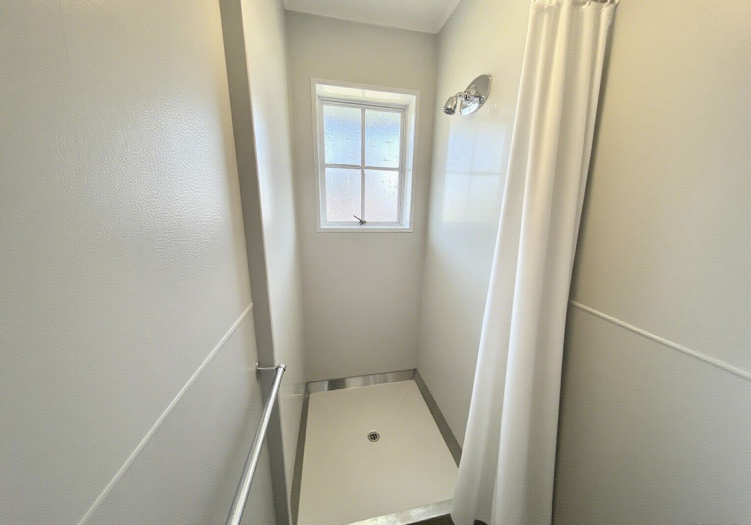 A narrow, white-walled bathroom with a shower, a window, and a door.