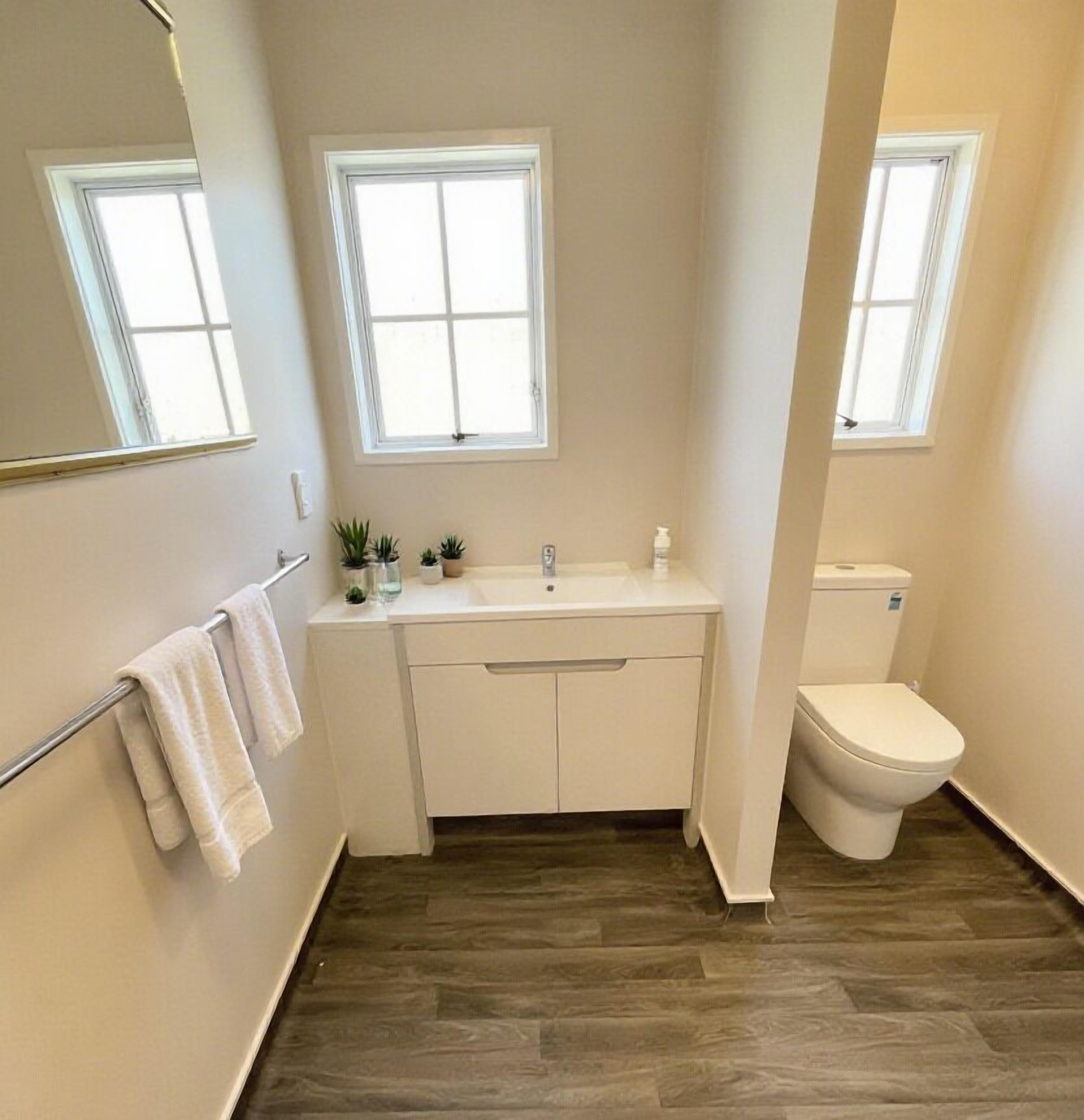 Un baño con un lavamanos blanco, un inodoro y dos ventanas.