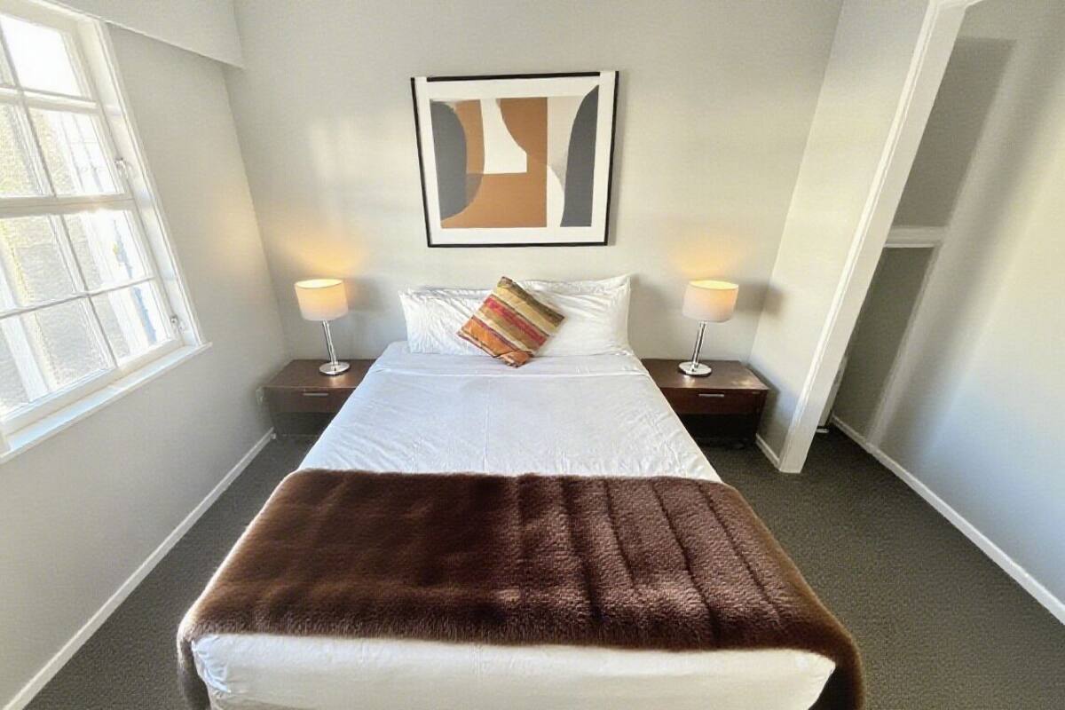 A neatly made bed with a brown throw blanket, two bedside lamps, a framed abstract artwork on the wall, and a window providing natural light.