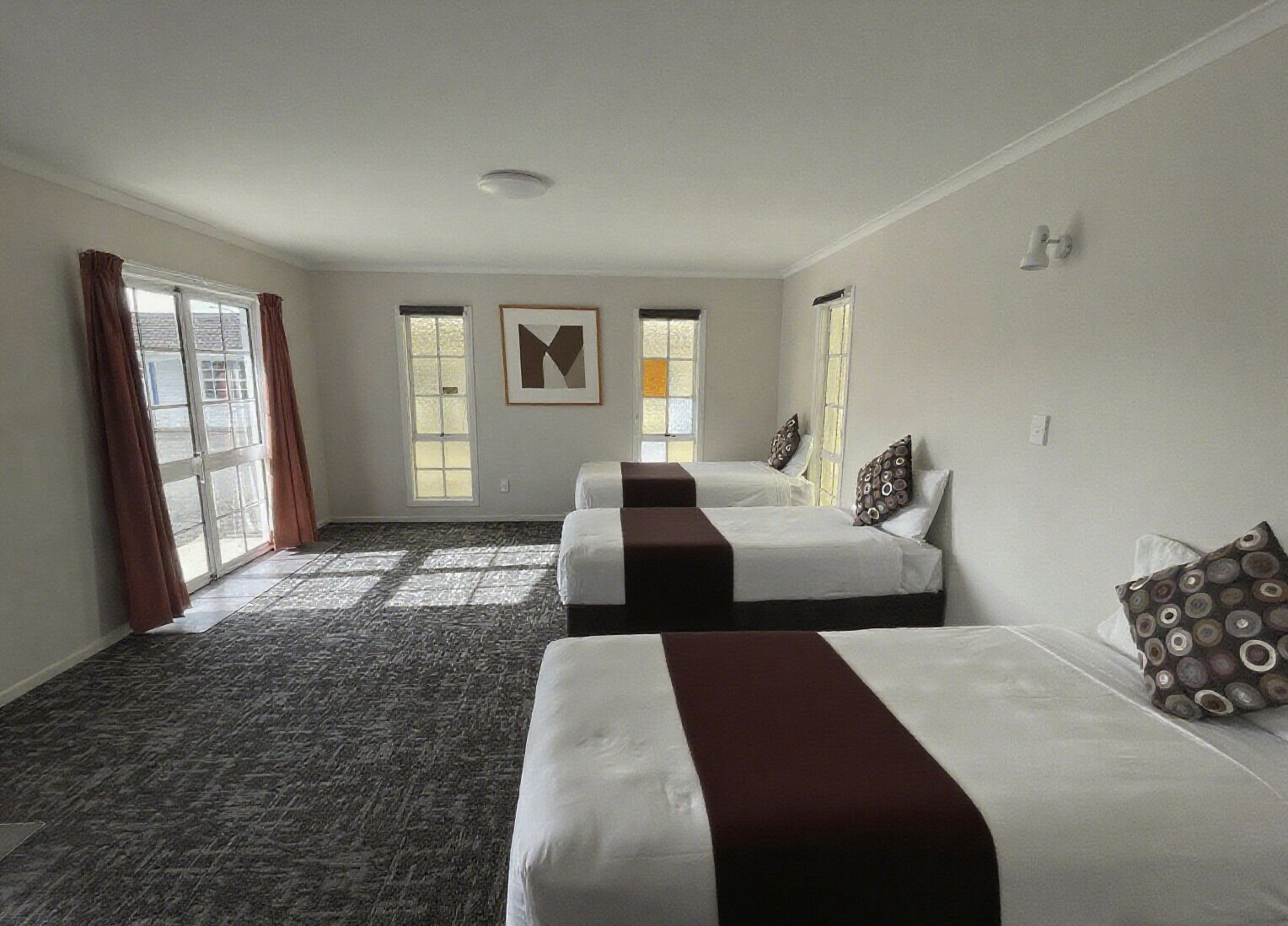 A hotel room with three beds, a large window with curtains, a patterned carpet, and a framed artwork on the wall.