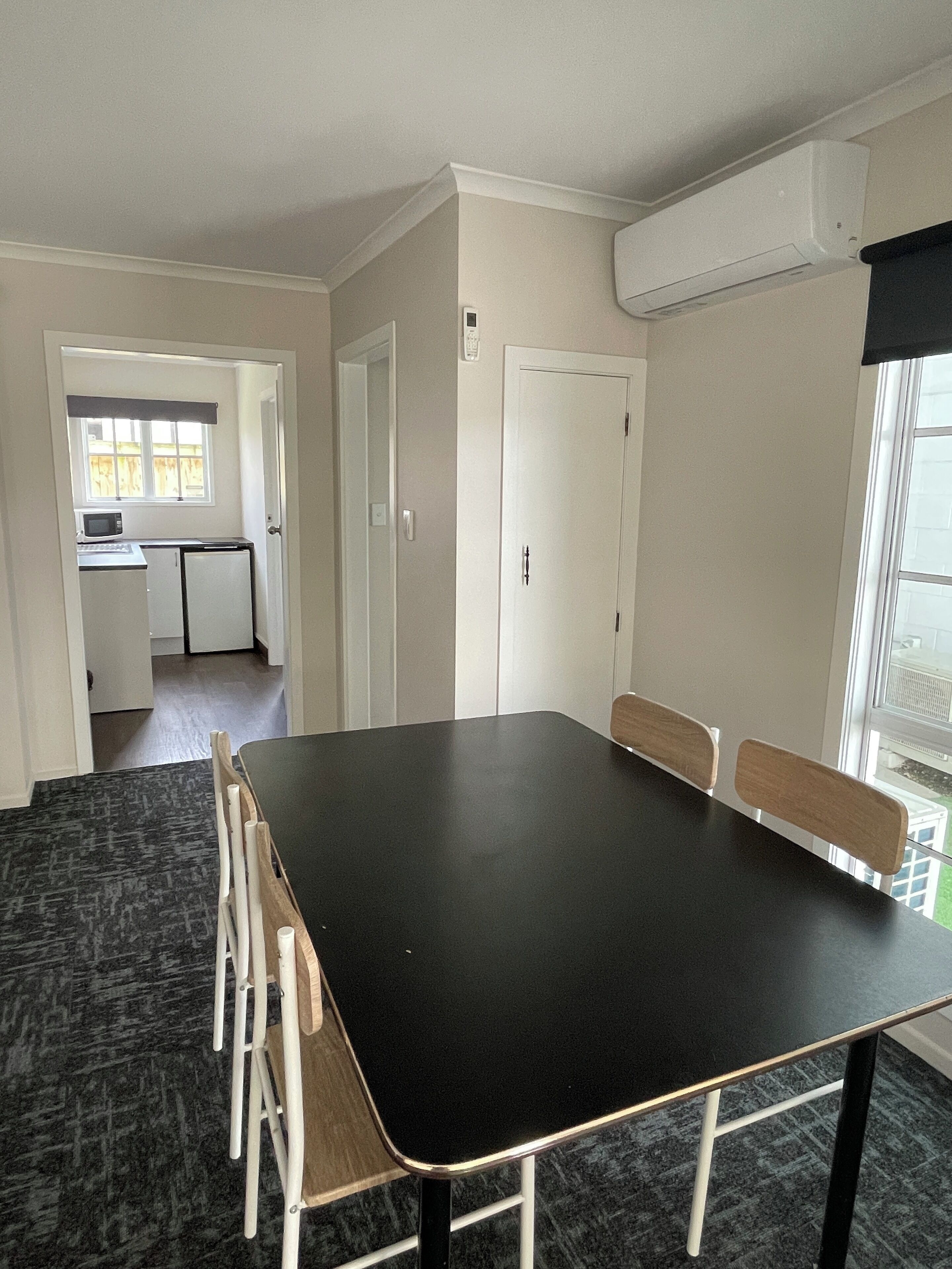 A room with a large black table, wooden chairs, a kitchen area with a microwave, and a window with a view.