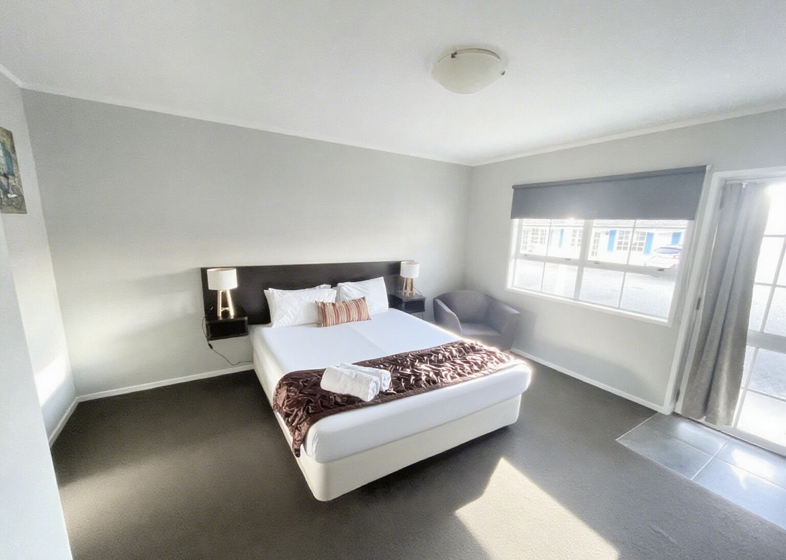 Una habitación de hotel moderna con una cama grande, una mesita de noche con lámpara, una silla y un ventanal con vistas a la calle.