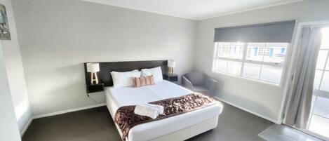 Una habitación de hotel moderna con una cama grande, una mesita de noche con lámpara, una silla y un ventanal con vistas a la calle.