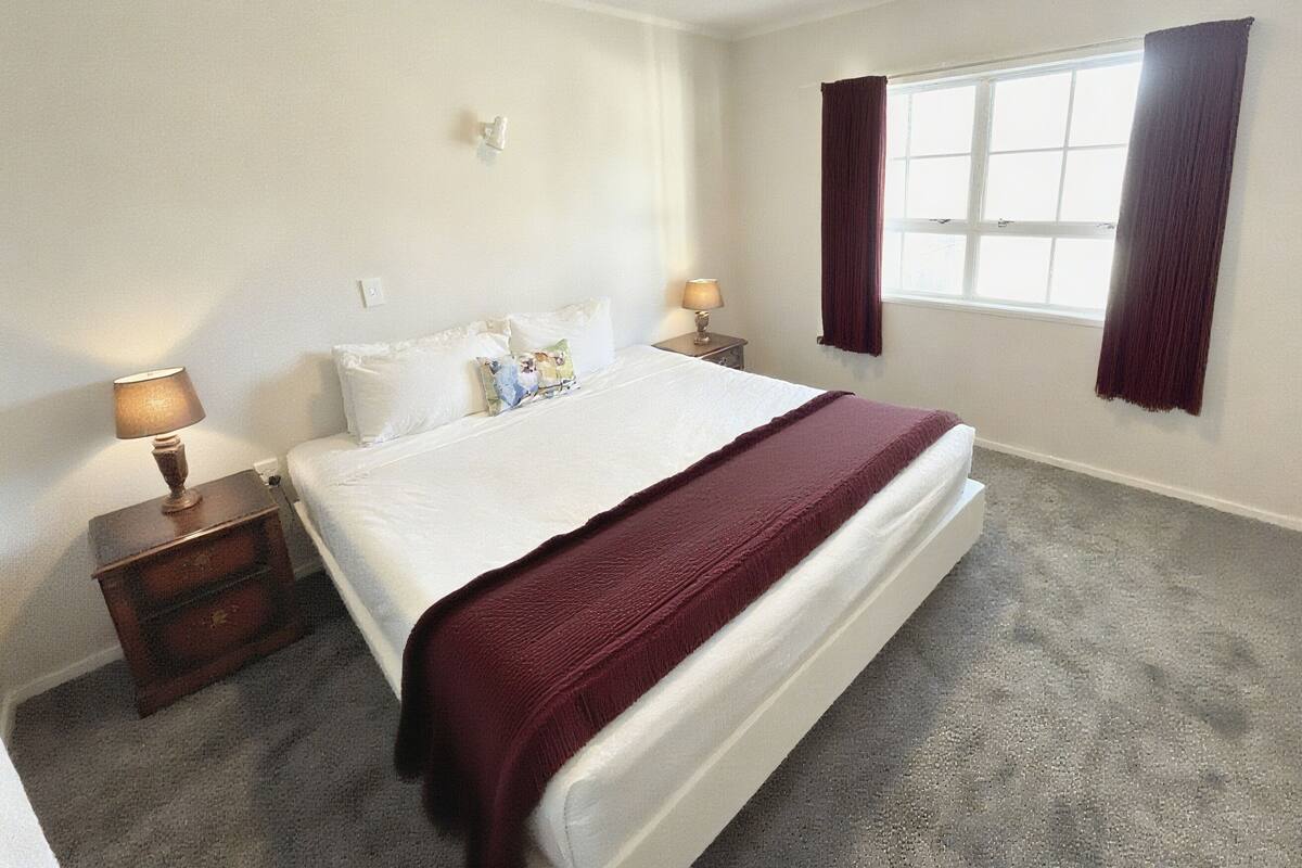 A bedroom with a large bed, two bedside tables, a window with curtains, and a wall-mounted light switch.