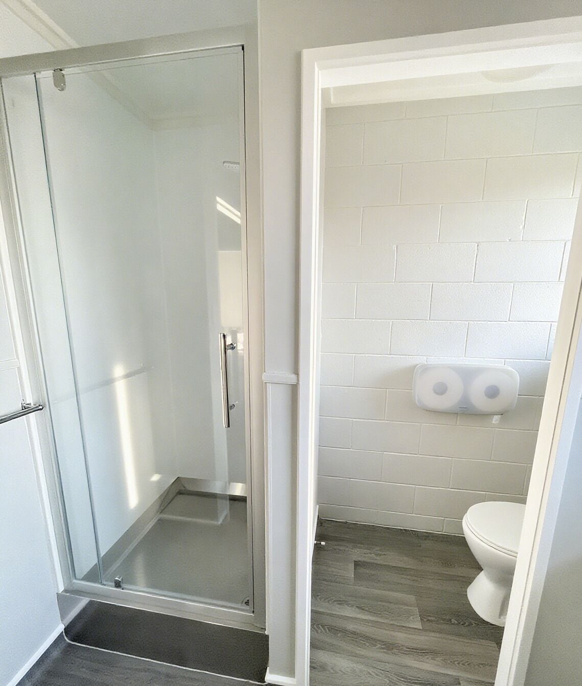 A bathroom with a glass shower door, white tiled walls, a toilet, and a roll of toilet paper on the wall.