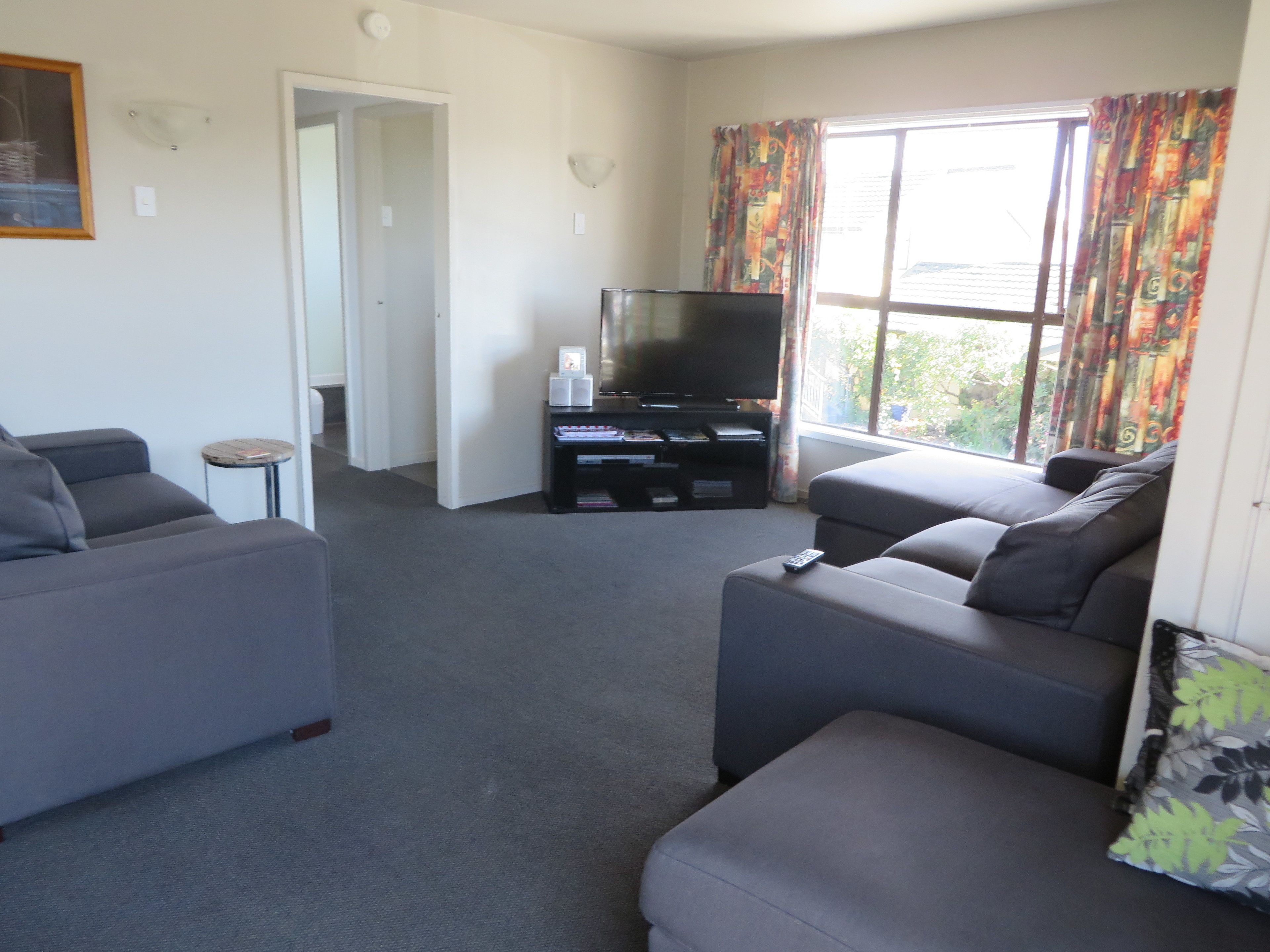 A living room with grey sofas, a TV on a black stand, and a window with colorful curtains.