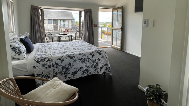 Un dormitorio con cama, una silla, un balcón con mesa y sillas, y vista a un edificio.