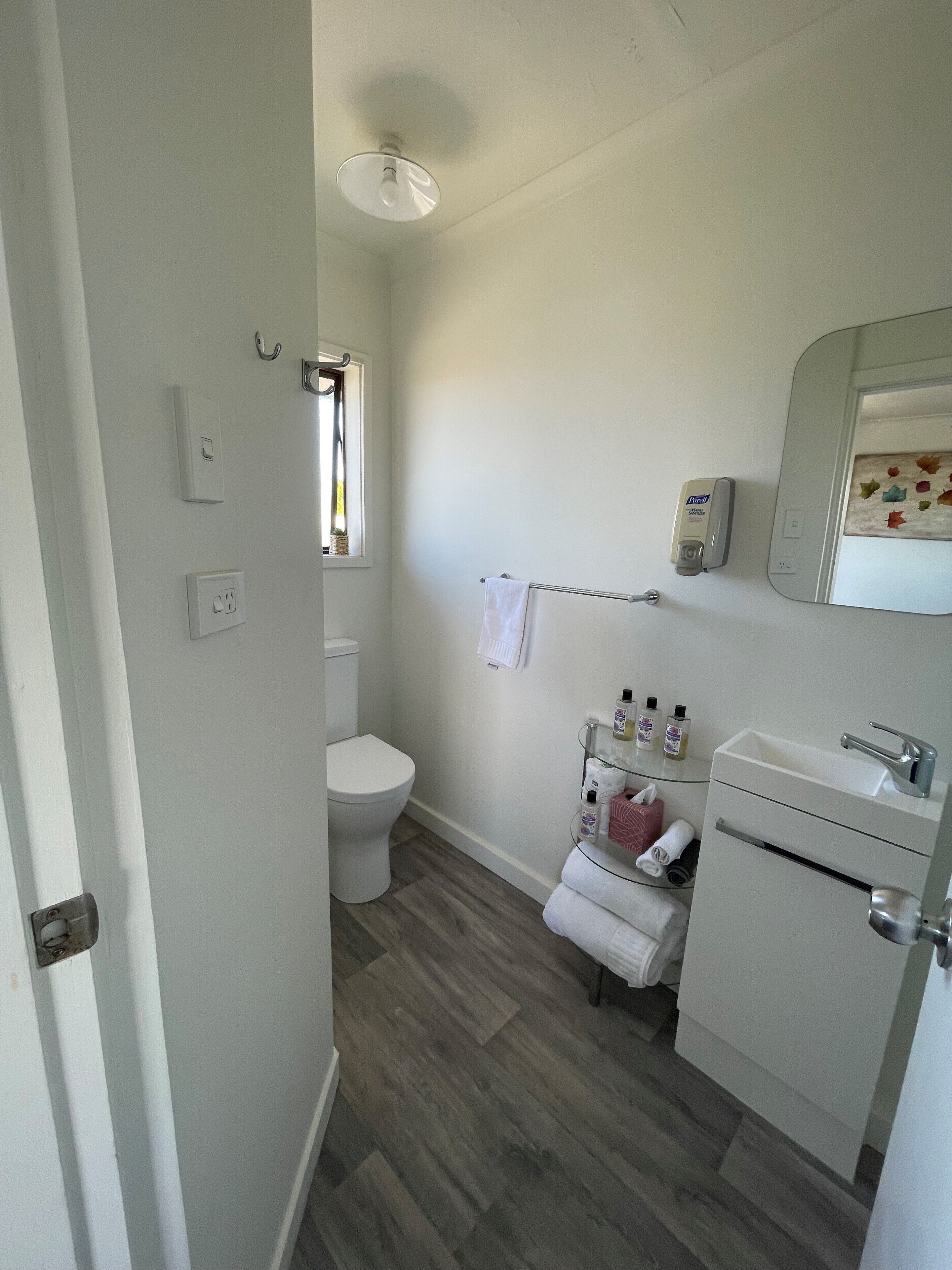 Un baño con inodoro, estante de vidrio con artículos de aseo, lavamanos y espejo.