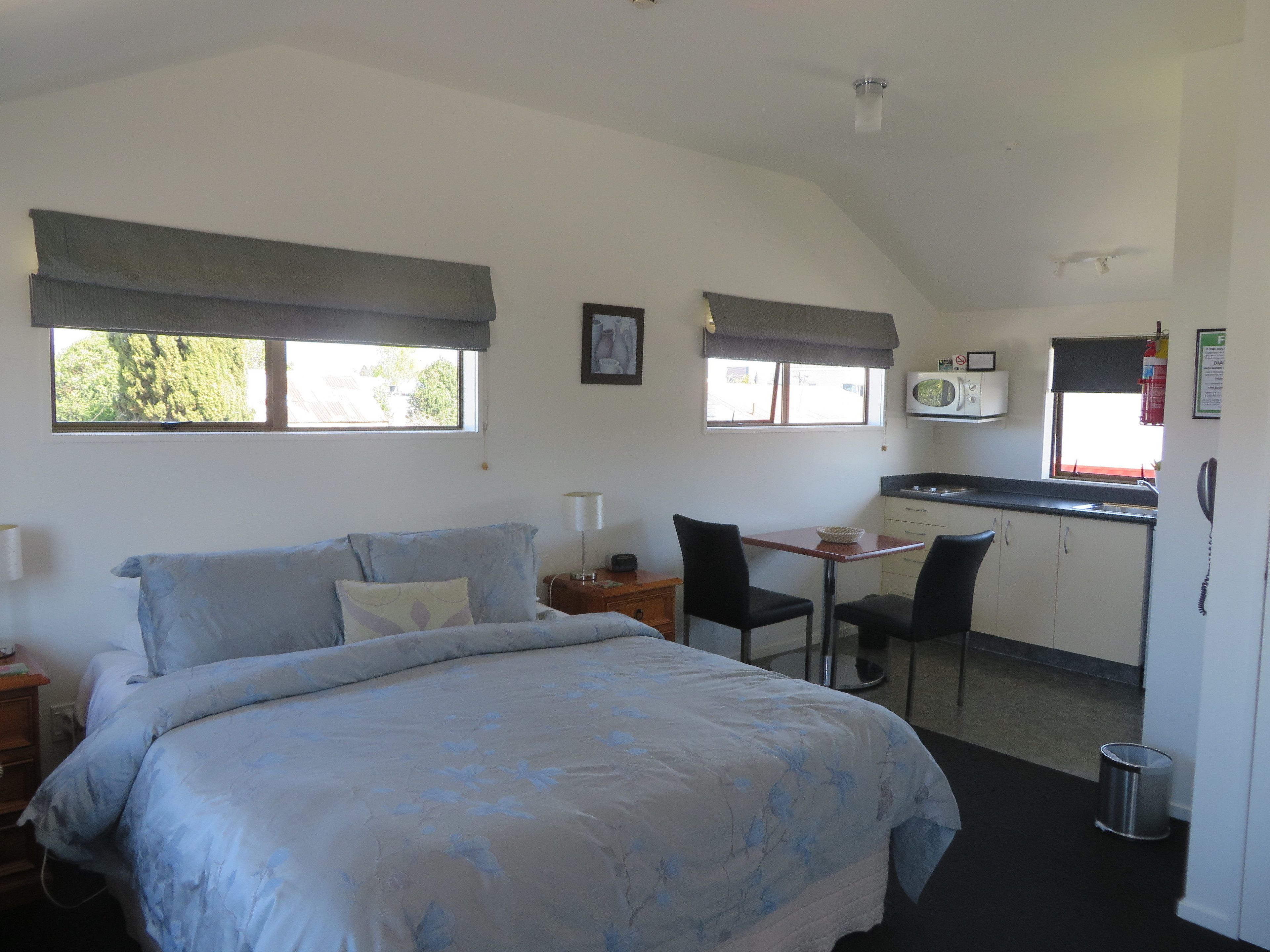 A bedroom with a bed, bedside tables, a desk, a chair, a microwave, and a small kitchenette.