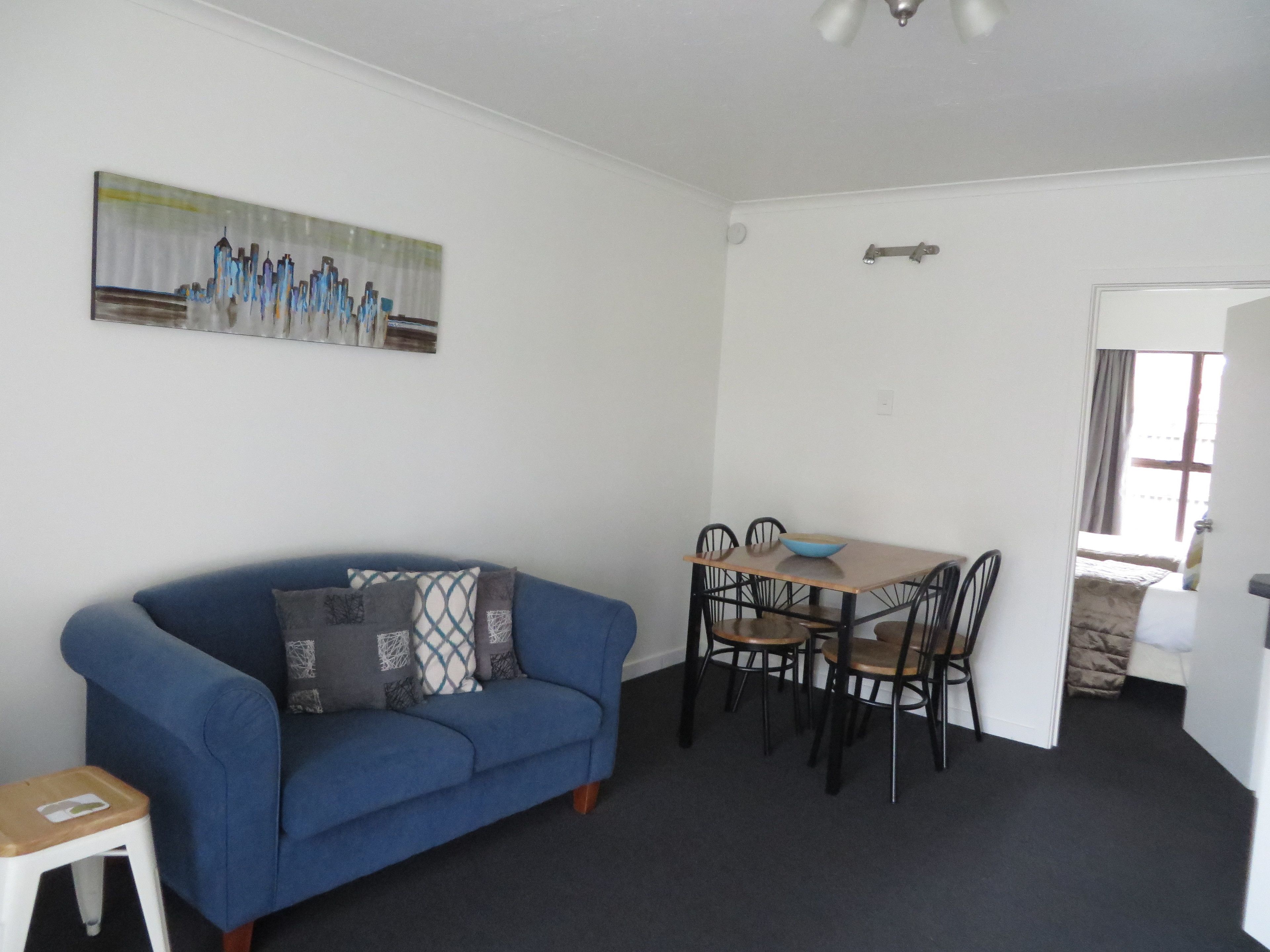 A room with a blue sofa, a dining table with four chairs, a painting on the wall, and a door leading to another room.