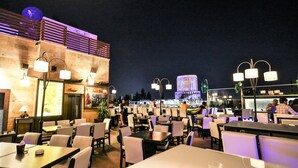 An outdoor dining area with tables and chairs, a bar area, and a view of a building with illuminated windows. - Hisham Hotel Amman (Amman)